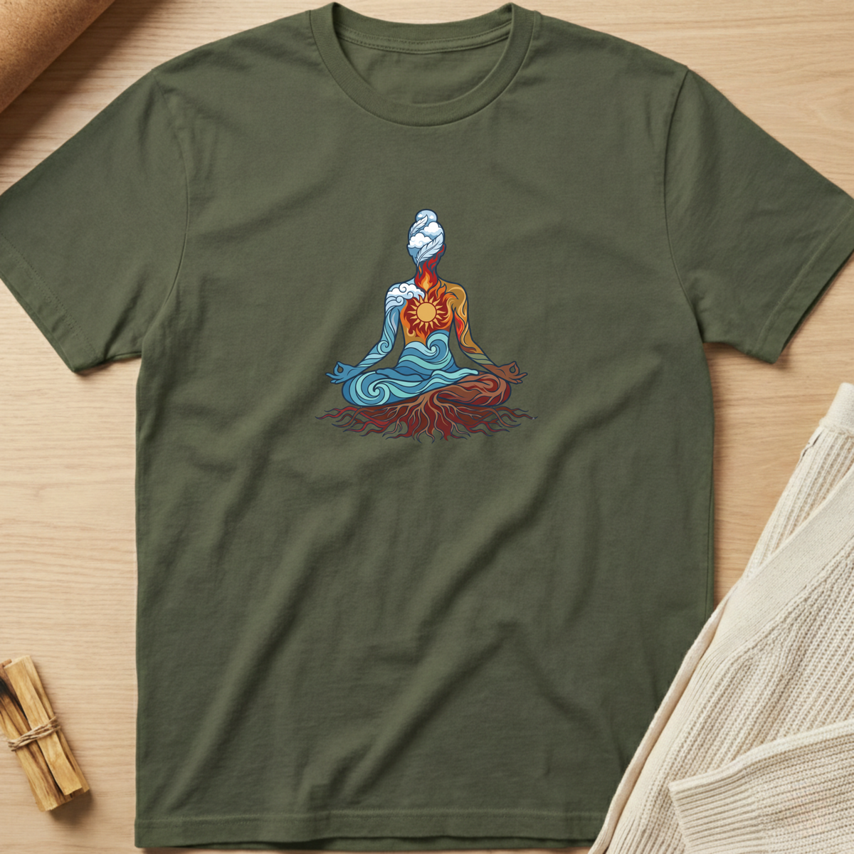 An olive green short-sleeve t-shirt with a colorful graphic of a meditating figure, laid flat on a light wood background with styled props. Minimalist yoga apparel by pemuyo.