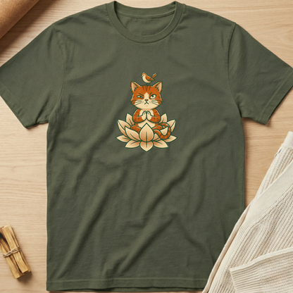 An olive green short-sleeve t-shirt laid flat on a light wood surface, featuring a central graphic of an orange cat meditating on a lotus flower with a small bird on its head. Minimalist yoga apparel by pemuyo.