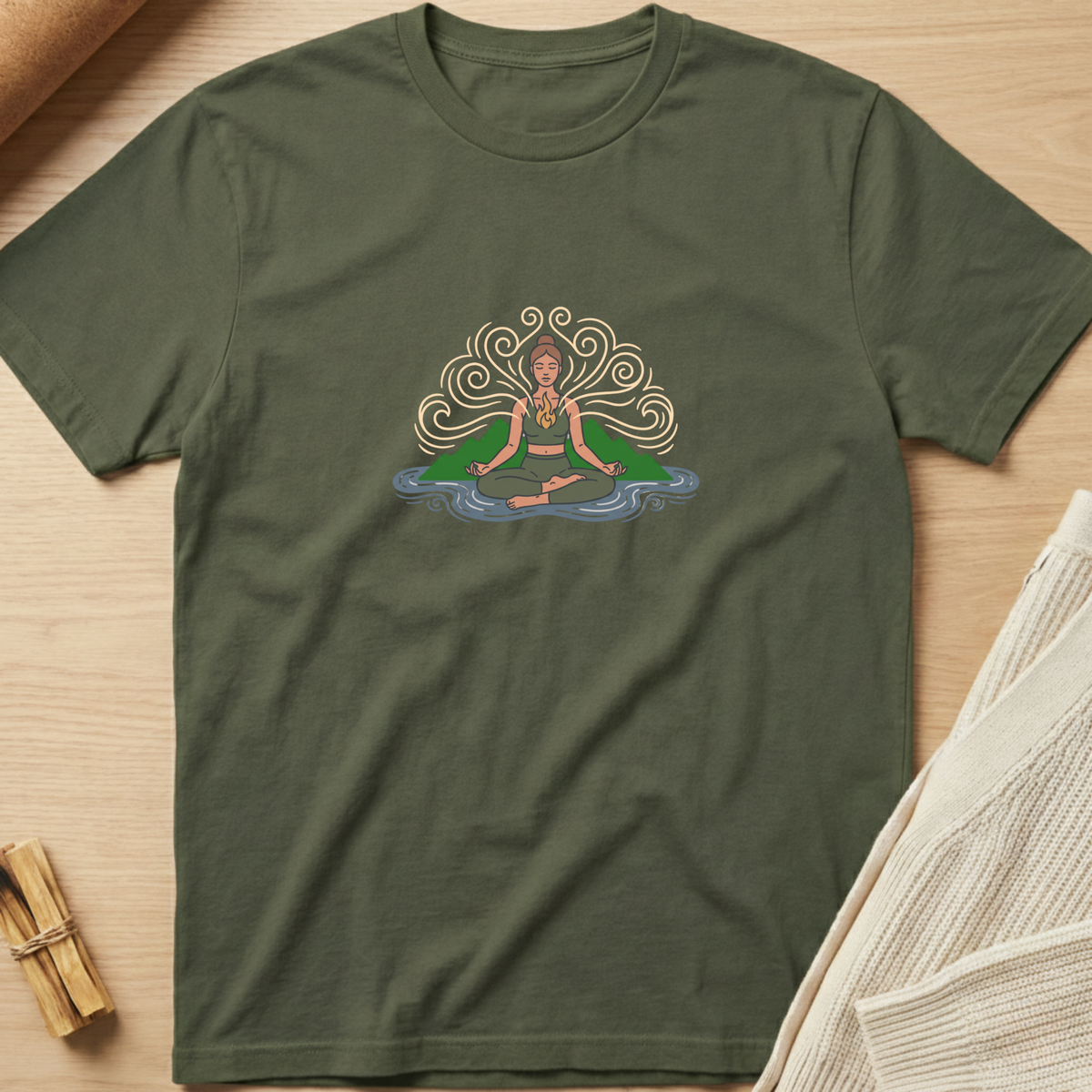 An olive green short-sleeve t-shirt with a colorful graphic of a meditating figure, laid flat on a light wood background with styled props. Minimalist yoga apparel by pemuyo.