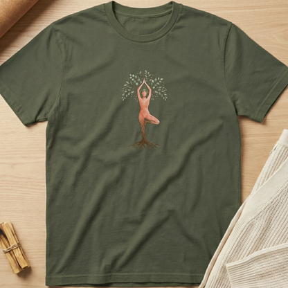 An olive green short-sleeve t-shirt laid flat on a light wood surface, featuring a central graphic of a tree with a person in a yoga pose growing from its trunk. The shirt is styled with a rolled brown paper towel and a beige knit fabric in the background. Minimalist yoga apparel by pemuyo.