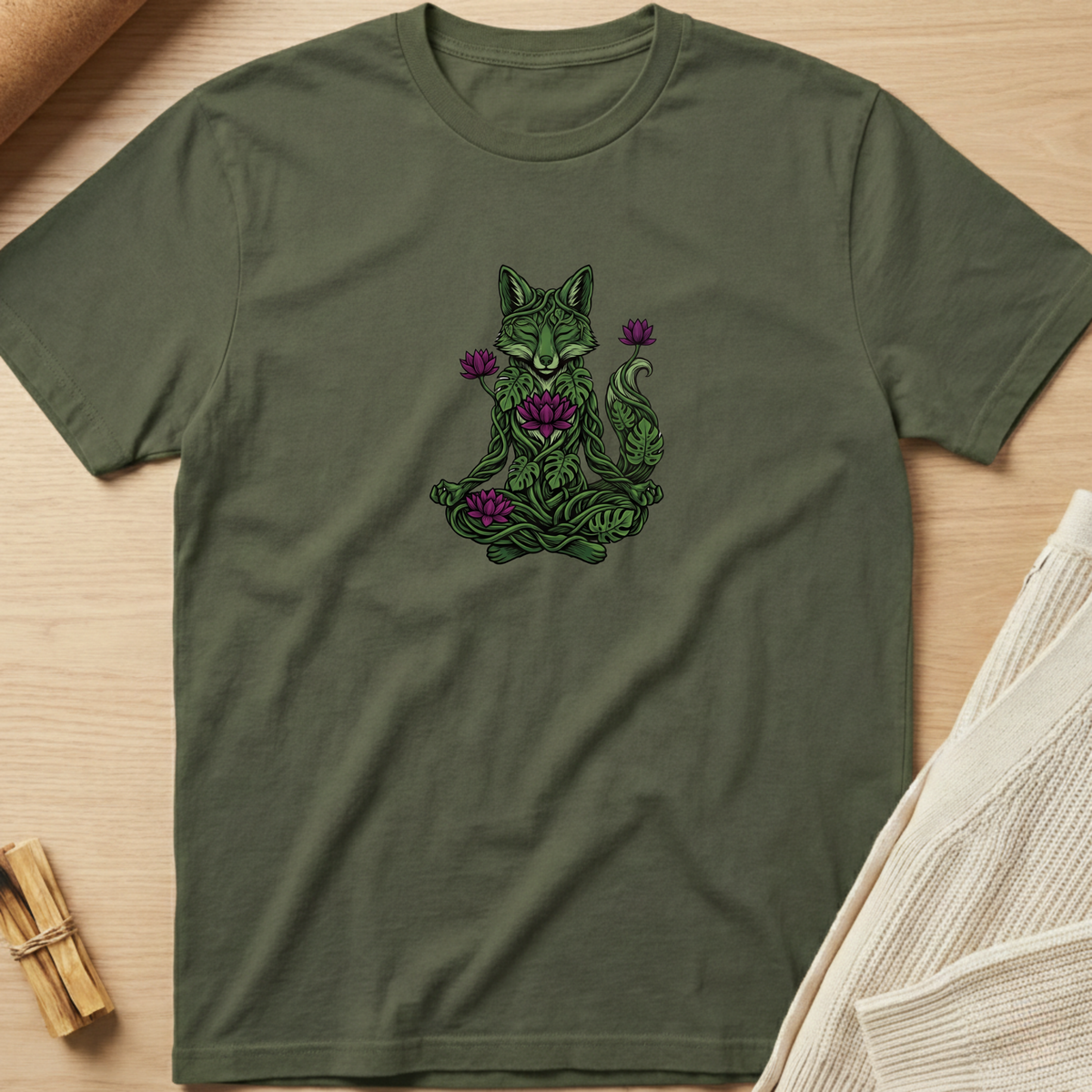 An olive green short-sleeve t-shirt with a black graphic of a meditating figure inside a tree, laid flat on a light wood background with styled props. Minimalist yoga apparel by pemuyo.