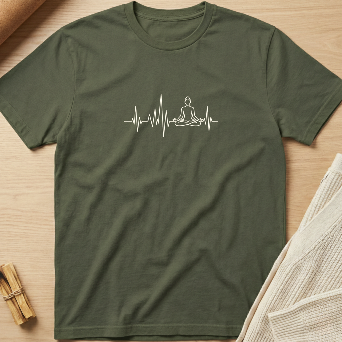 An olive green short-sleeve t-shirt with a white graphic of a meditating figure on an EKG line, laid flat on a light wood background with styled props. Minimalist yoga apparel by pemuyo.