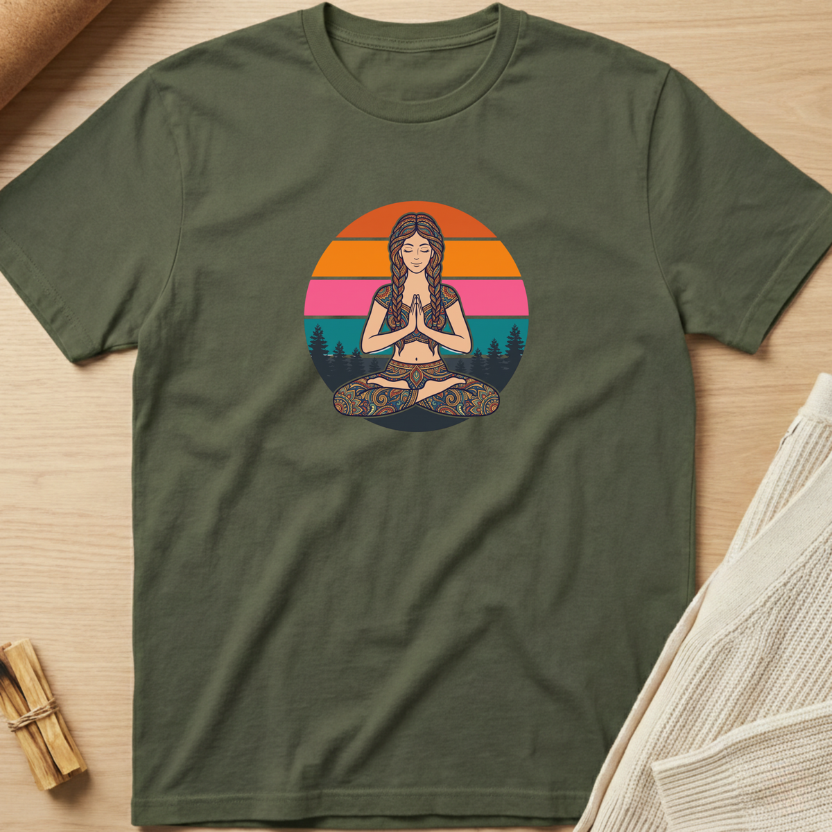 An olive green short-sleeve t-shirt with a colorful graphic of a woman meditating in front of a sunset circle, laid flat on a light wood background with a small bundle of incense and a folded cream knit blanket nearby. Minimalist yoga apparel by pemuyo.