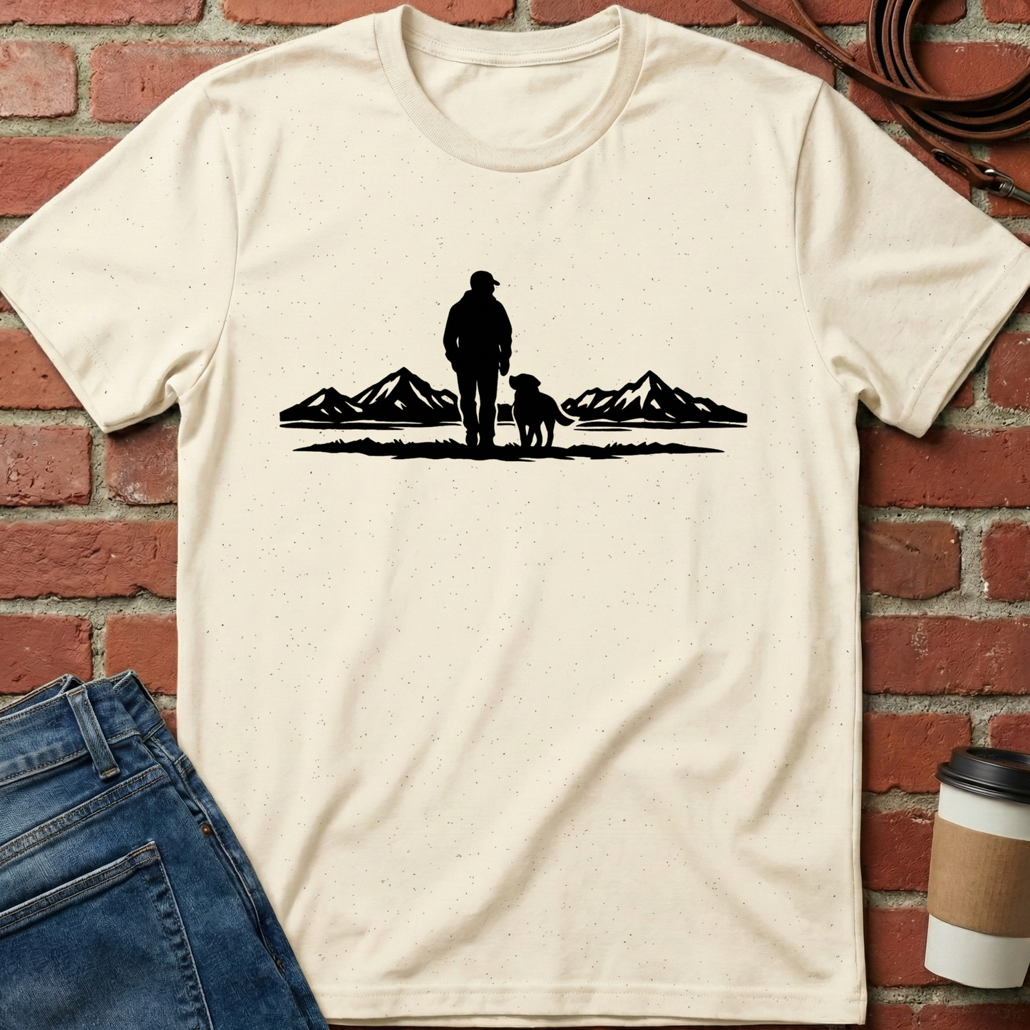 Beige t-shirt with a black graphic of a person and dog standing in front of mountains, placed on a brick surface with jeans and a coffee cup. Minimalist dog apparel by pemuyo.