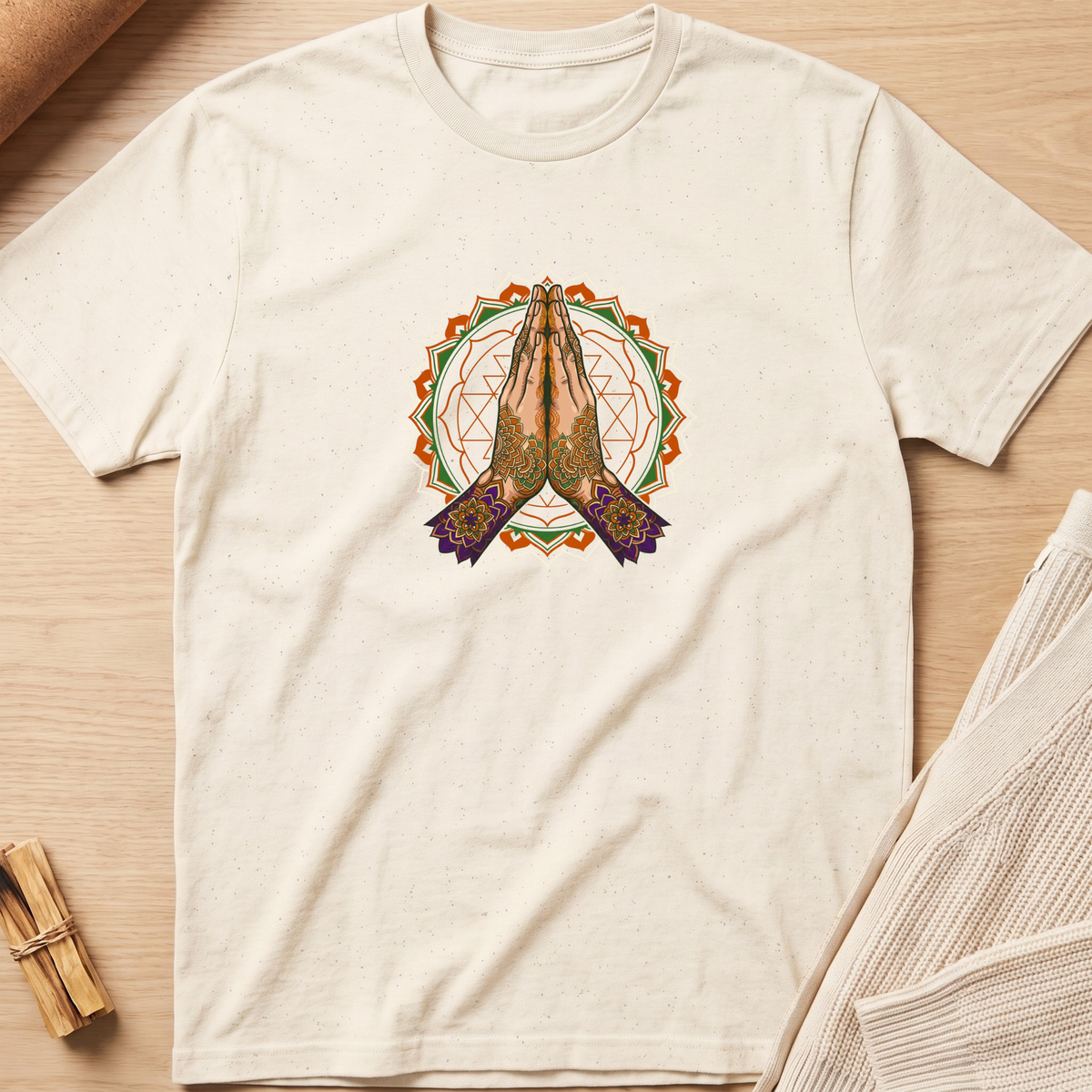 An off-white short-sleeve t-shirt with a central graphic of praying hands inside a geometric mandala design, laid flat on a light wood background with cozy props. Minimalist yoga apparel by pemuyo.