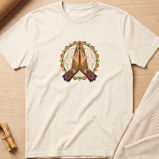 An off-white short-sleeve t-shirt with a central graphic of praying hands inside a geometric mandala design, laid flat on a light wood background with cozy props. Minimalist yoga apparel by pemuyo.