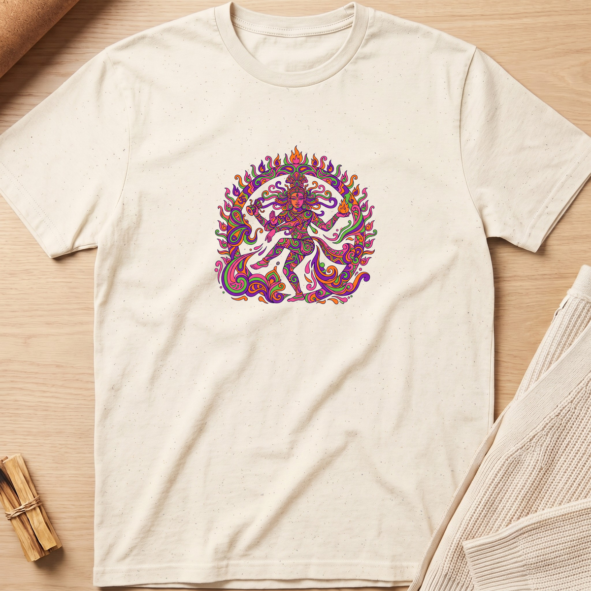 An off-white crewneck t-shirt featuring a colorful, detailed graphic of a dancing Hindu deity (Shiva Nataraja) in the center, laid flat on a light wood background with styled props. Minimalist yoga apparel by pemuyo.