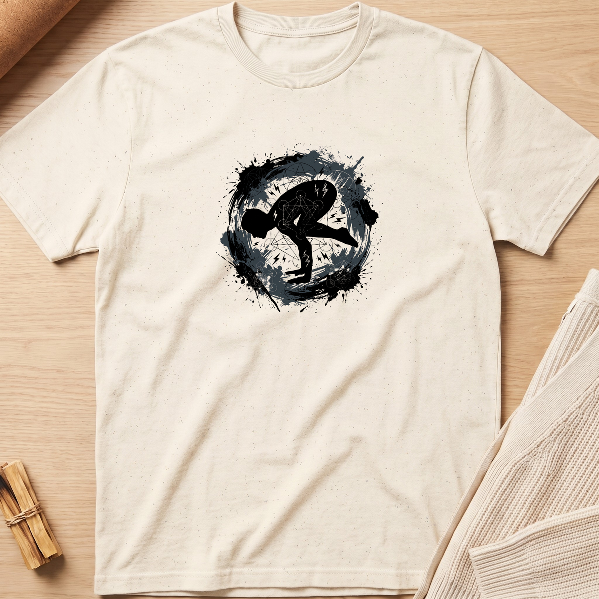 An off-white short-sleeve t-shirt laid flat on a light wood background, featuring a black circular graphic with a stylized human silhouette in the center. Minimalist yoga apparel by pemuyo.