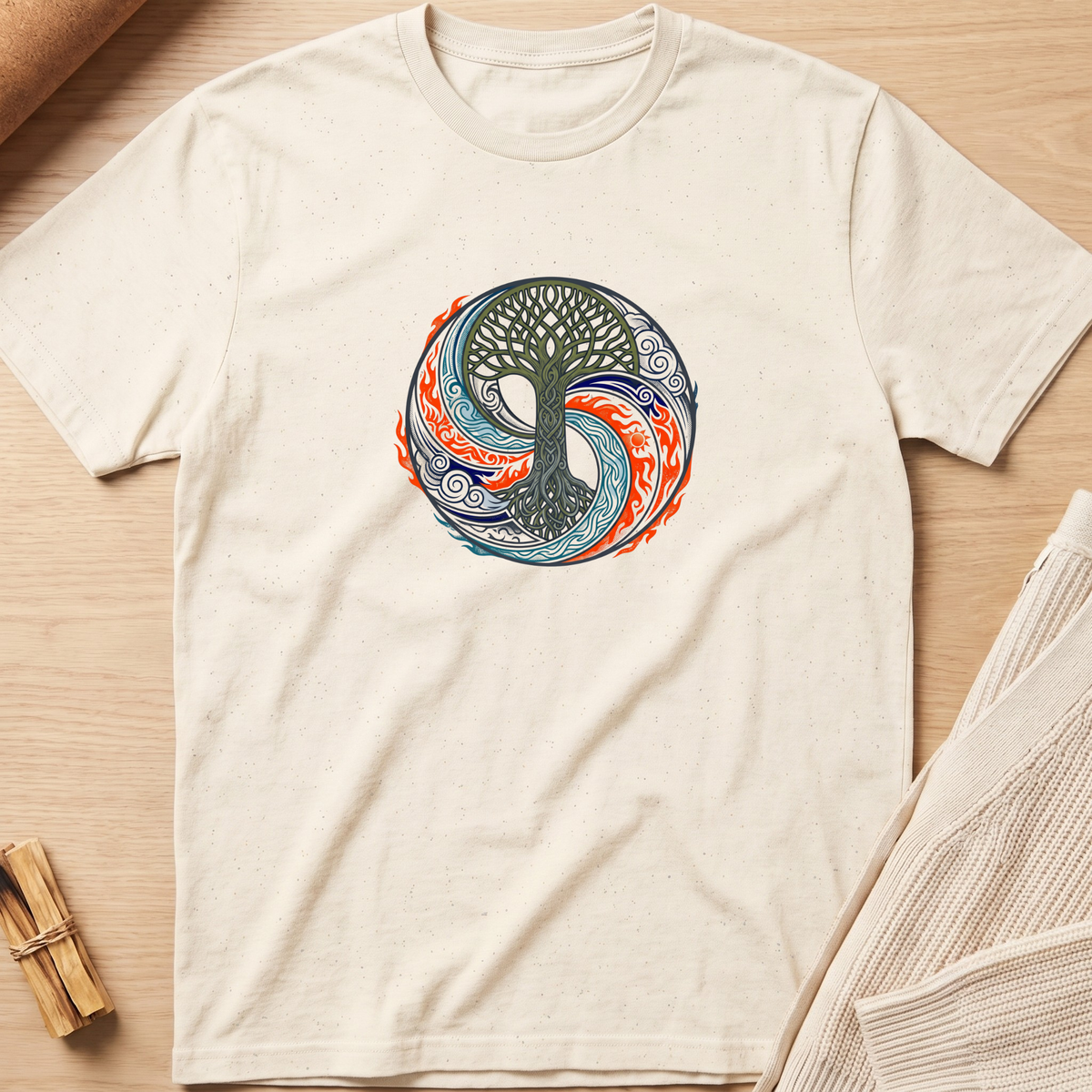 An off-white short-sleeve t-shirt with a circular 'Tree of Life' yin-yang style graphic in blue, orange, and green, laid flat on a light wood background with a knit blanket and small wooden sticks nearby. Minimalist yoga apparel by pemuyo.