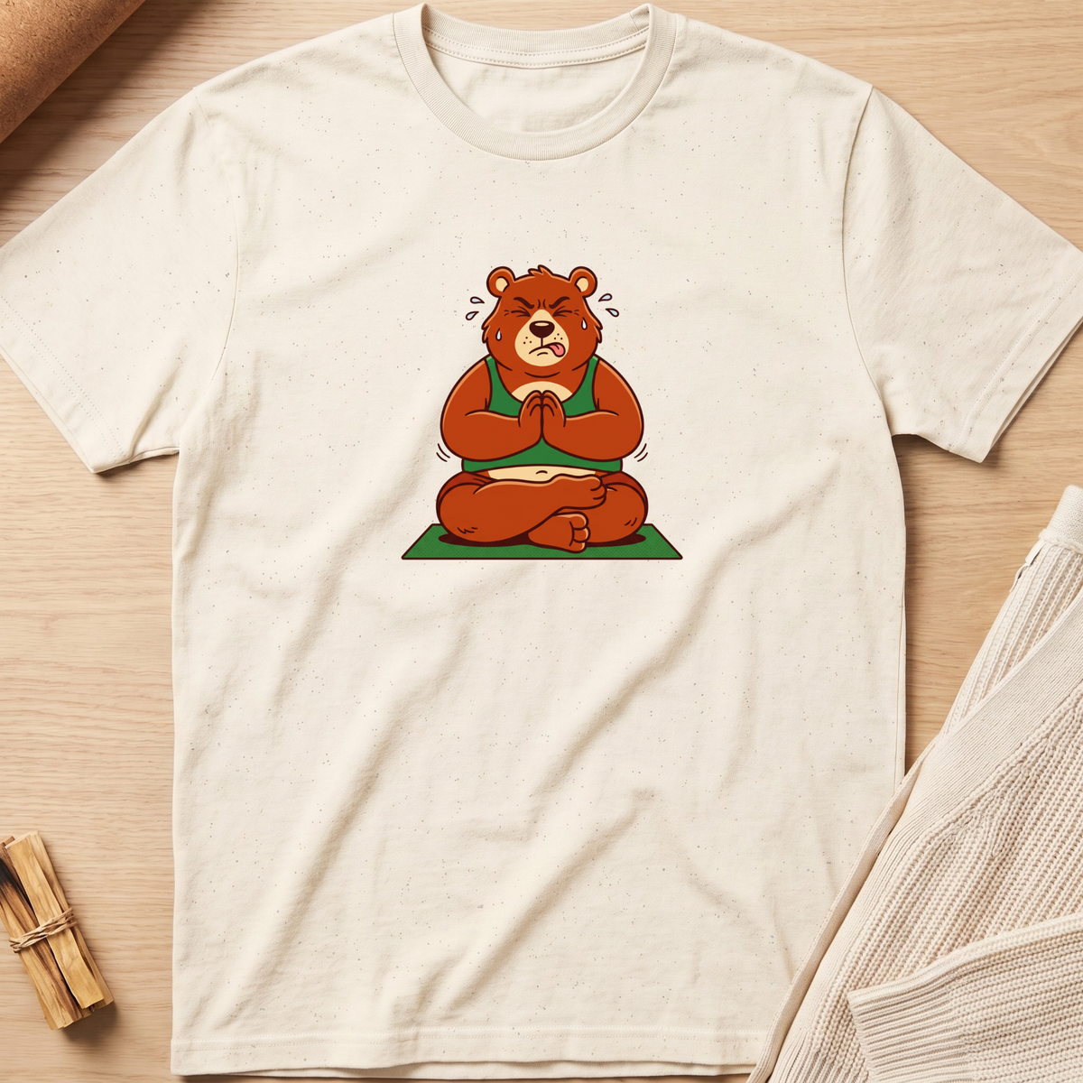 An off-white short-sleeve t-shirt with a graphic of a brown bear meditating in a green robe, laid flat on a light wood background with a knit sweater and wooden sticks as props. Minimalist yoga apparel by pemuyo.
