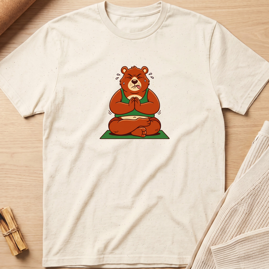An off-white short-sleeve t-shirt with a graphic of a brown bear meditating in a green robe, laid flat on a light wood background with a knit sweater and wooden sticks as props. Minimalist yoga apparel by pemuyo.