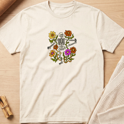 An off-white short-sleeve t-shirt with a colorful graphic of a skeleton surrounded by flowers, laid flat on a light wood background. Minimalist yoga apparel by pemuyo.