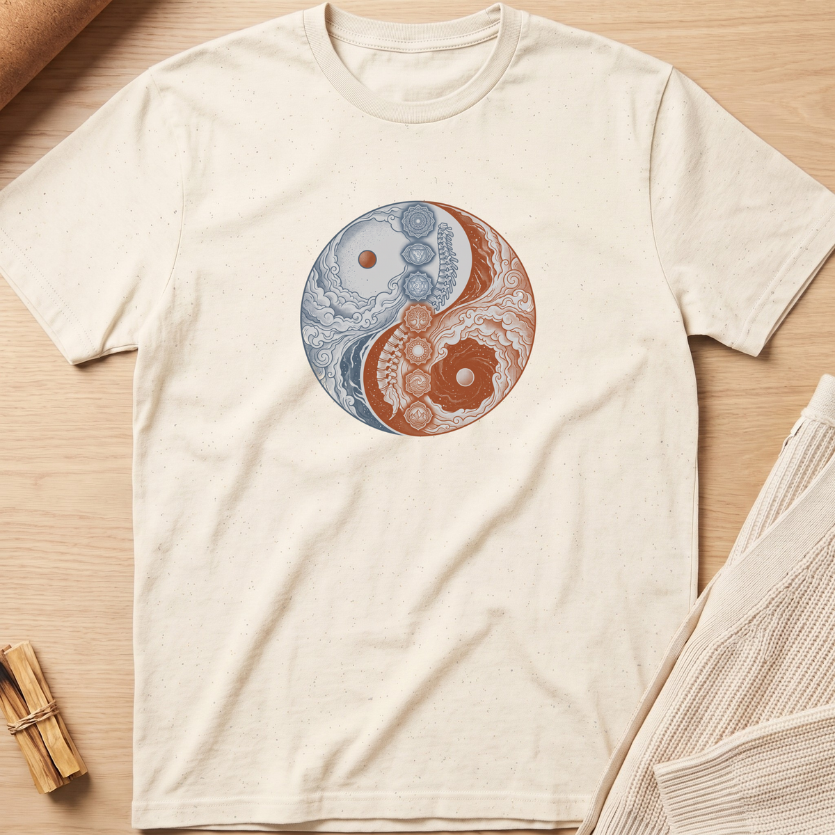 An off-white short-sleeve t-shirt laid flat on a light wood surface, featuring a circular yin-yang style graphic on the chest with blue and rust-orange ornamental details. Minimalist yoga apparel by pemuyo.