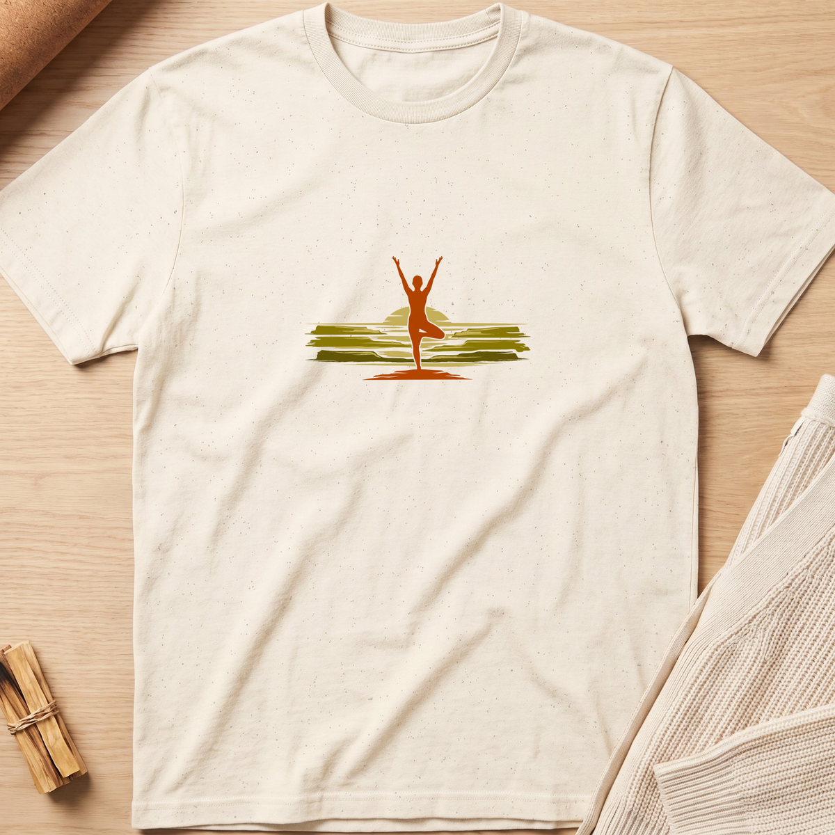 An off-white short-sleeve t-shirt with a small graphic of a yoga pose against a sunset on the chest, laid flat on a light wood background with styled props. Minimalist yoga apparel by pemuyo.