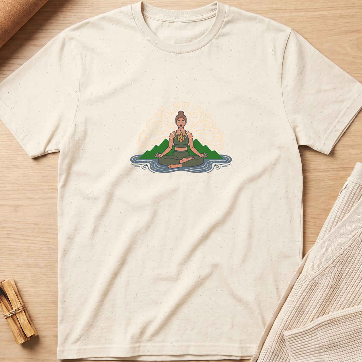 An off-white short-sleeve t-shirt with a colorful graphic of a woman meditating on a lotus flower, laid flat on a light wood background. Minimalist yoga apparel by pemuyo.