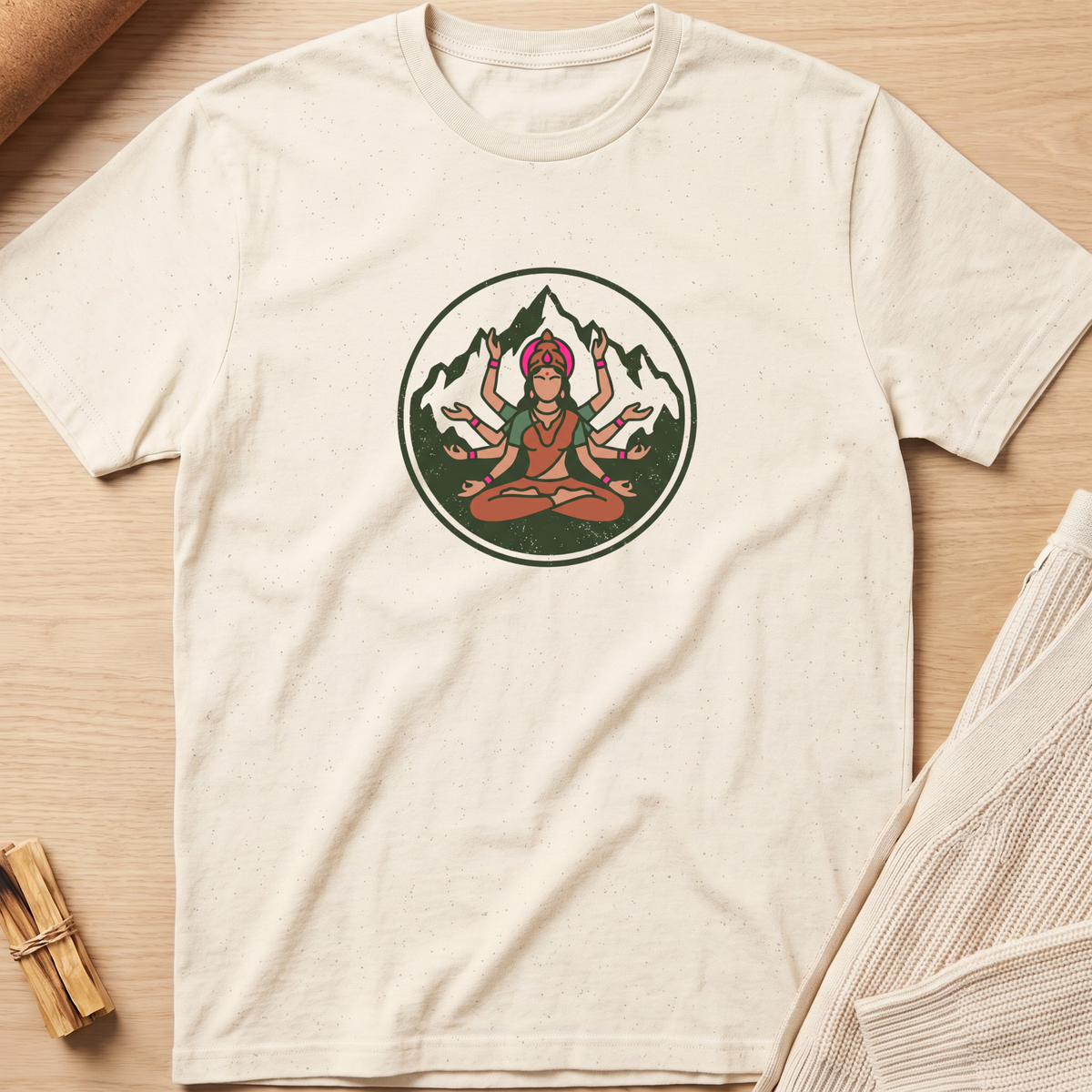 An off-white short-sleeve t-shirt with a circular graphic of a multi-armed deity meditating in front of mountains, laid flat on a light wood background with styled props. Minimalist yoga apparel by pemuyo.