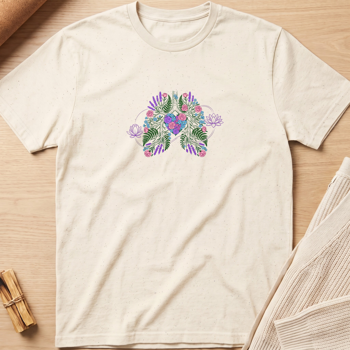 An off-white short-sleeve t-shirt with a colorful tropical floral and leaf graphic on the chest, laid flat on a light wood background. Minimalist yoga apparel by pemuyo.