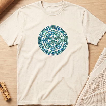 An off-white short-sleeve t-shirt with a blue and green geometric Sri Mandala print on the chest, laid flat on a light wood background with styled props. Minimalist yoga apparel by pemuyo.