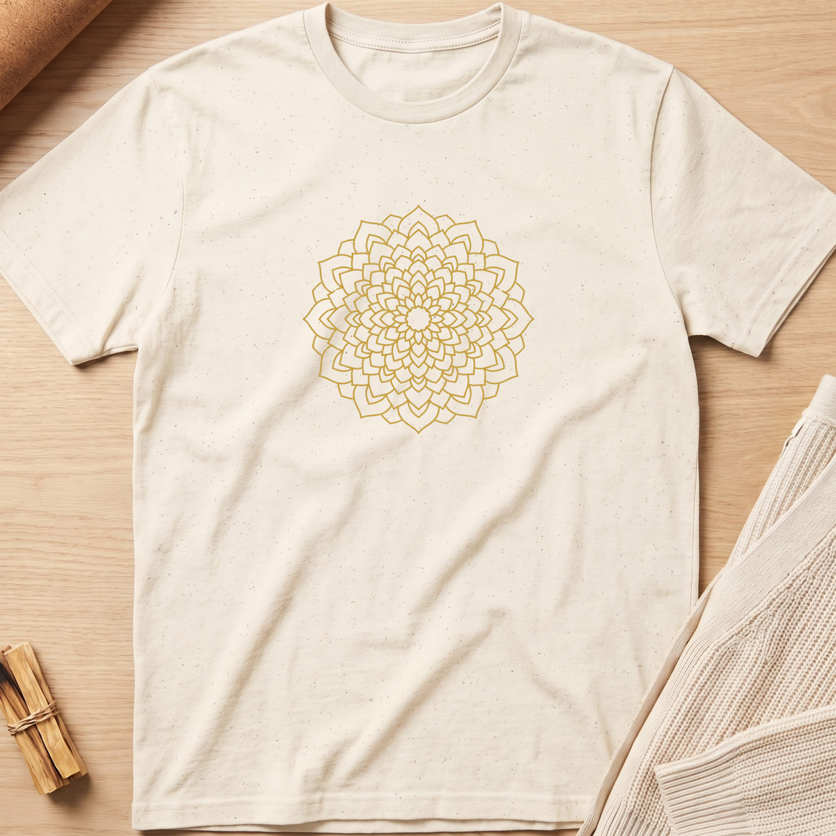 An off-white short-sleeve t-shirt with a gold mandala graphic printed on the chest, laid flat on a light wood background with neutral props. Minimalist yoga apparel by pemuyo.