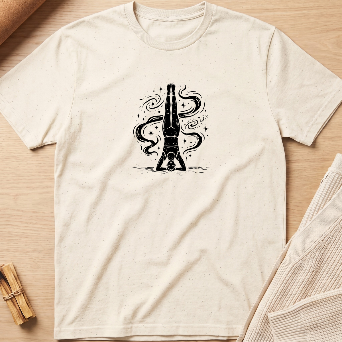 An off-white short-sleeve t-shirt with a black graphic of a bottle balanced on its cap, surrounded by swirling smoke, laid flat on a light wood background. Minimalist yoga apparel by pemuyo.