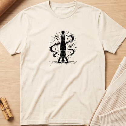 An off-white short-sleeve t-shirt with a black graphic of a bottle balanced on its cap, surrounded by swirling smoke, laid flat on a light wood background. Minimalist yoga apparel by pemuyo.
