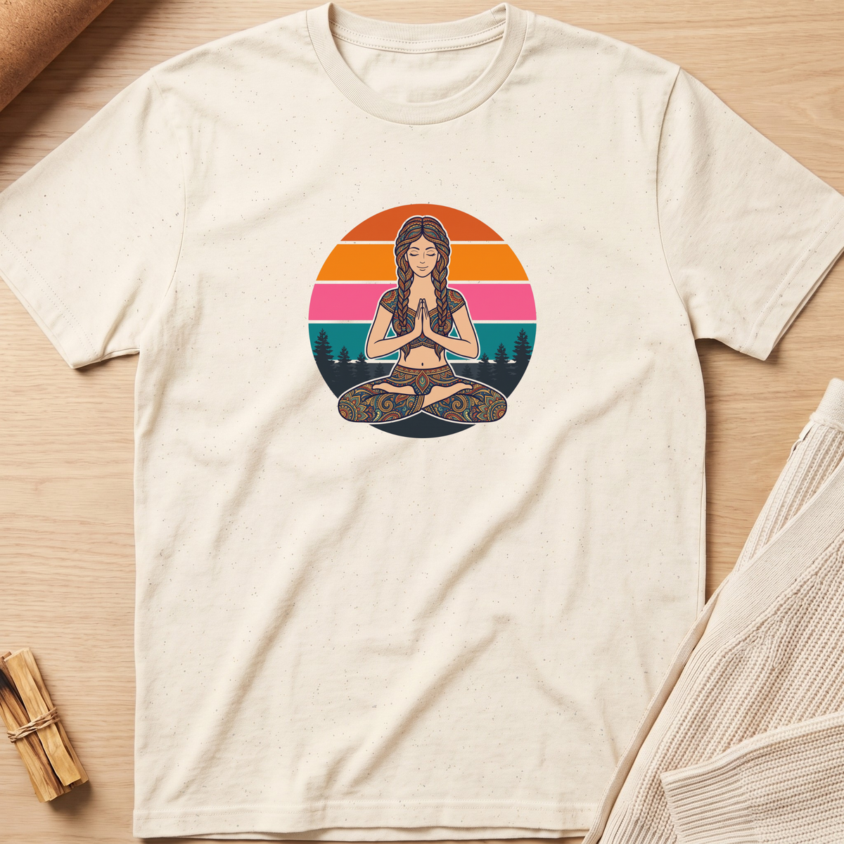 An off-white short-sleeve t-shirt with a colorful graphic of a woman meditating in a lotus position, set against a circular sunset background. The shirt is laid flat on a light wood surface with neutral props. Minimalist yoga apparel by pemuyo.