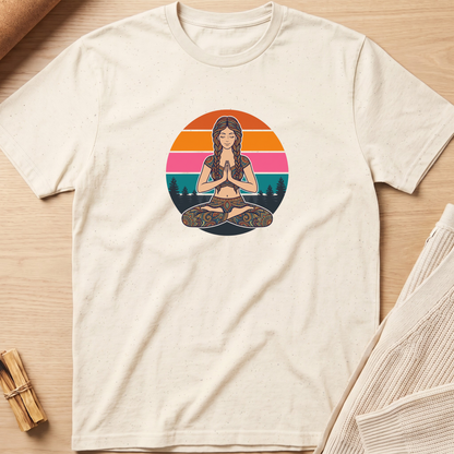 An off-white short-sleeve t-shirt with a colorful graphic of a woman meditating in a lotus position, set against a circular sunset background. The shirt is laid flat on a light wood surface with neutral props. Minimalist yoga apparel by pemuyo.