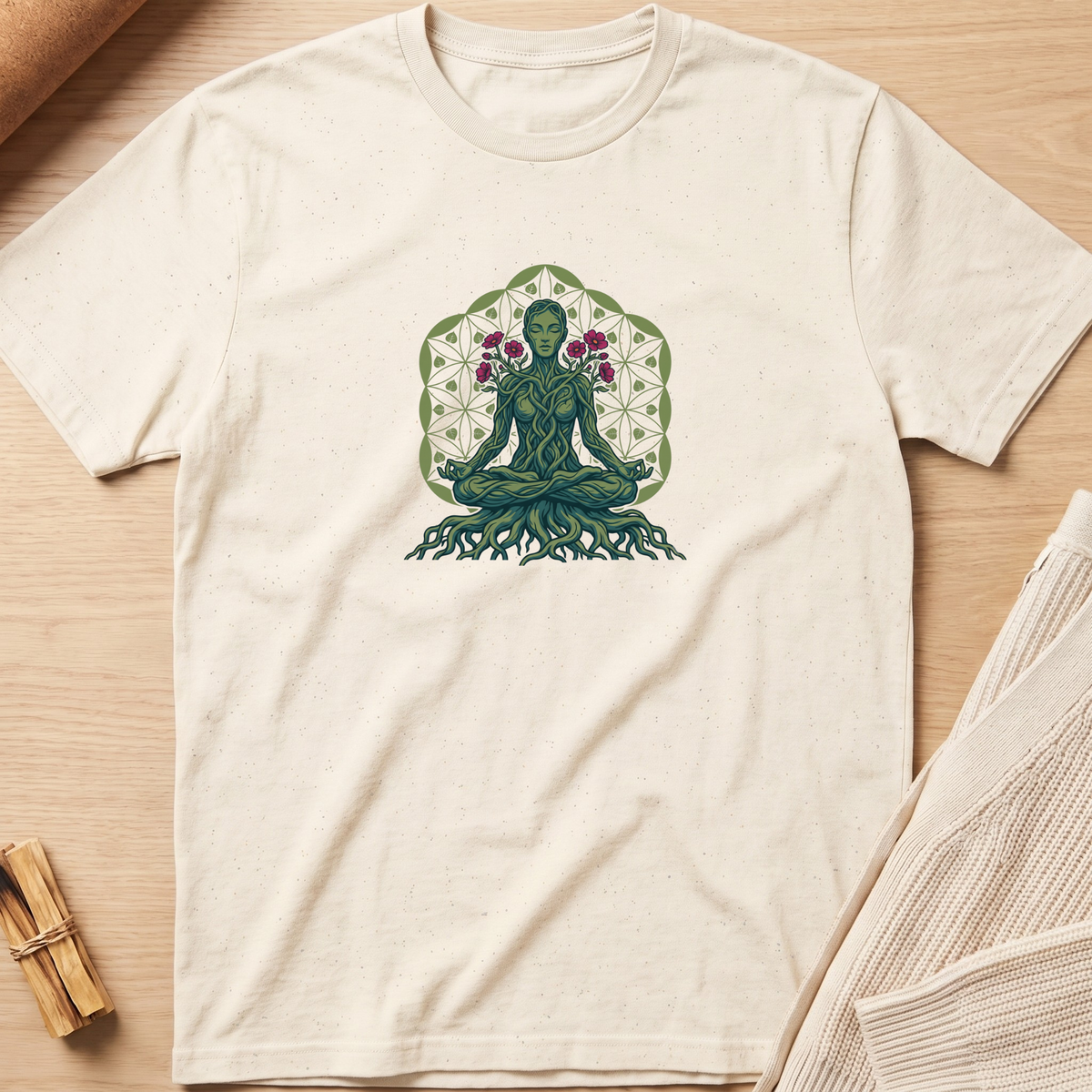 An off-white short-sleeve t-shirt laid flat on a light wood background, featuring a central graphic of a green meditating figure with roots and flowers inside a geometric pattern. Minimalist yoga apparel by pemuyo.
