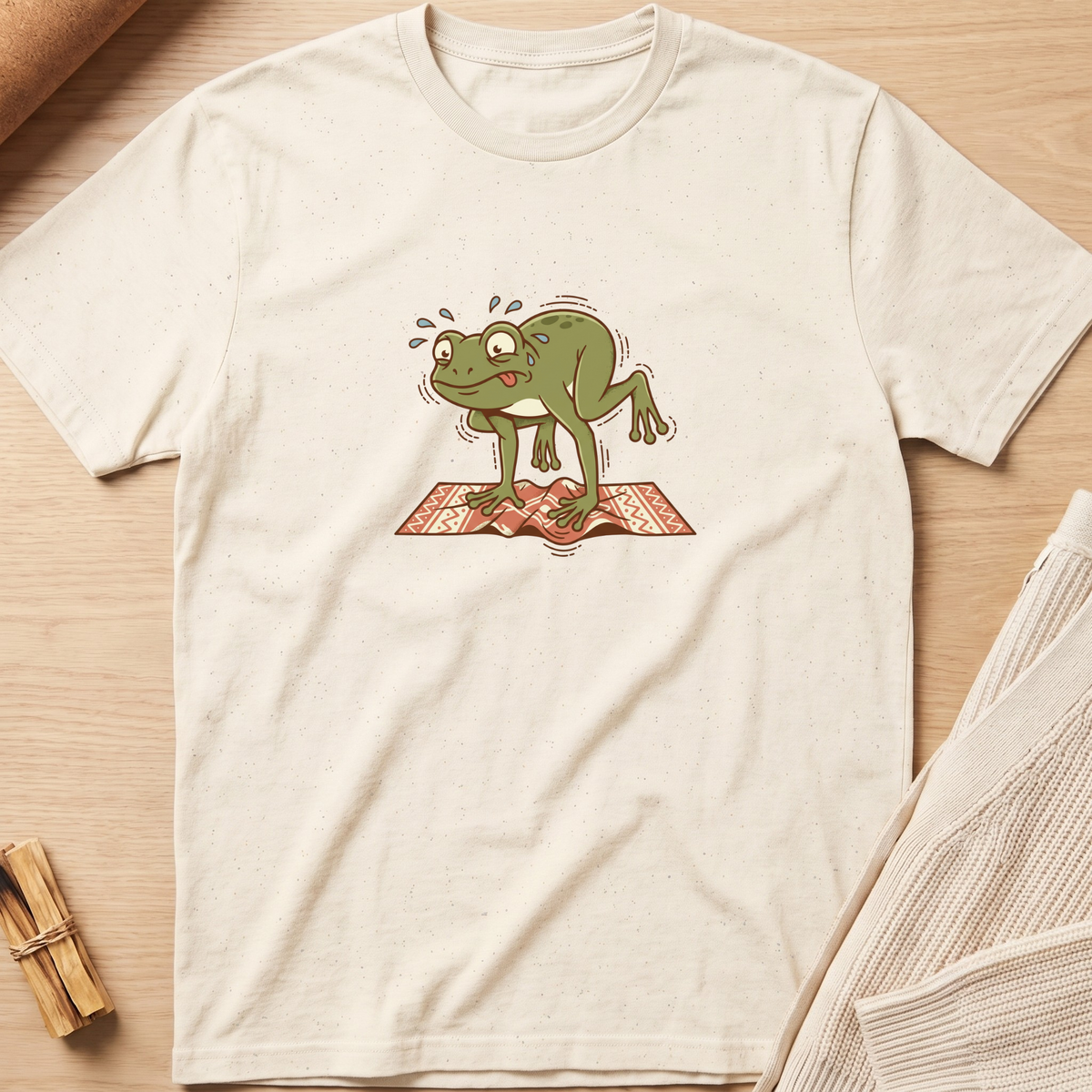 An off-white short-sleeve t-shirt with a graphic of a sweating green frog jumping on a red patterned rug, laid flat on a light wood background with styled props. Minimalist yoga apparel by pemuyo.