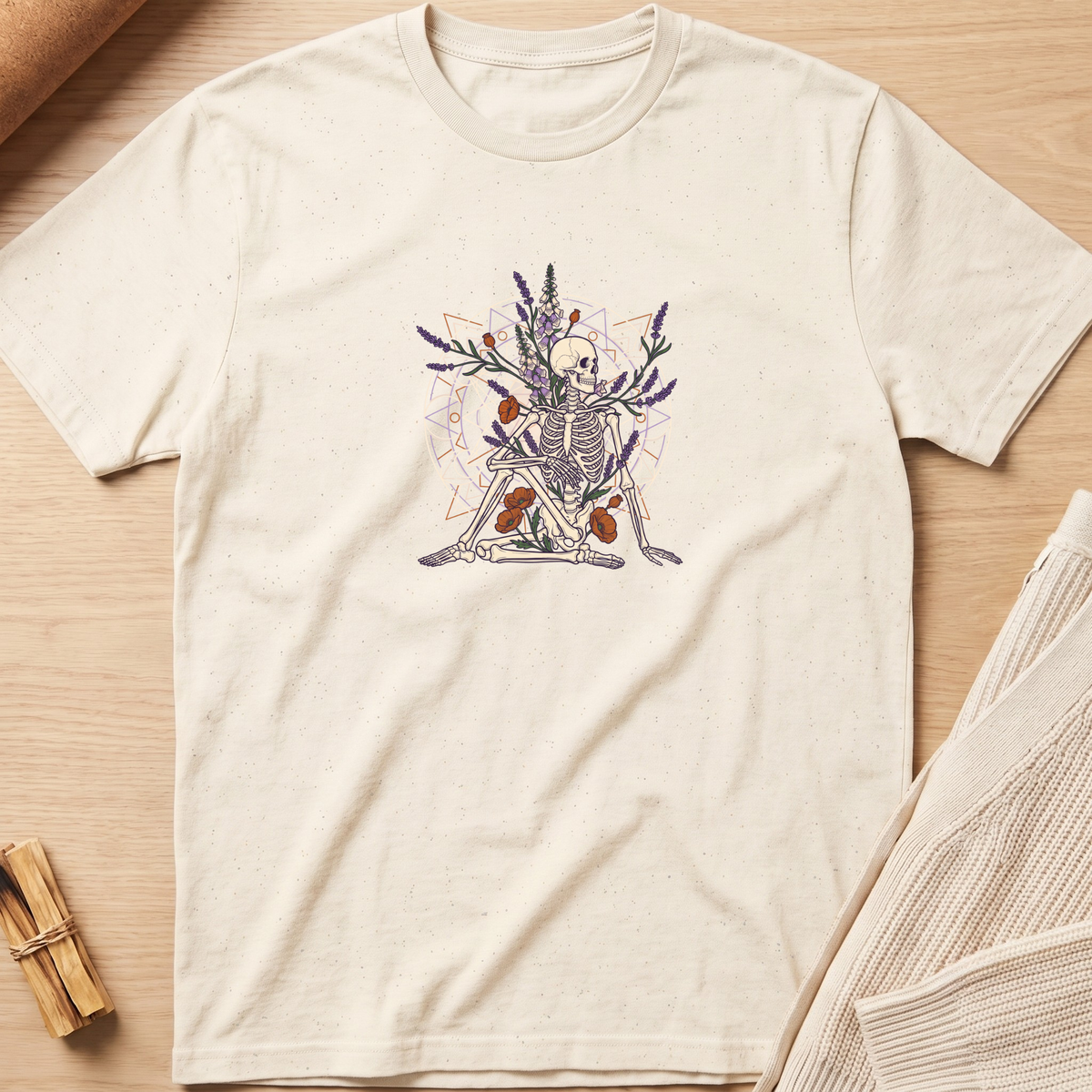 An off-white short-sleeve t-shirt with a central graphic of a seated skeleton surrounded by purple lavender, pinecones, and geometric shapes, laid flat on a light wood background. Minimalist yoga apparel by pemuyo.