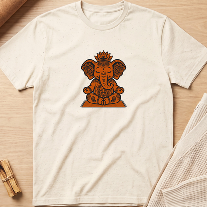 An off-white short-sleeve t-shirt with a central graphic of Ganesha in orange and brown tones, laid flat on a light wood background with neutral props. Minimalist yoga apparel by pemuyo.