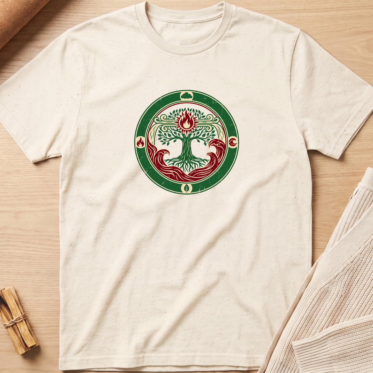 An off-white short-sleeve t-shirt laid flat on a light wood background, featuring a circular green and red tree of life graphic printed on the chest. Minimalist yoga apparel by pemuyo.