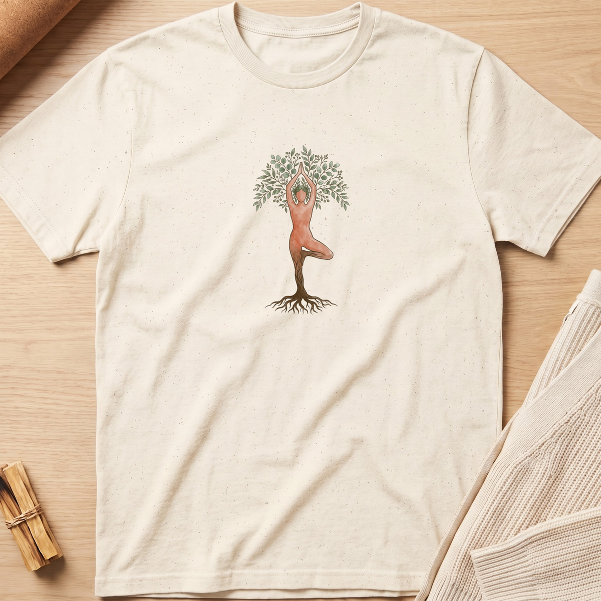 An off-white short-sleeve t-shirt with a central graphic of a tree trunk with roots and a figure in a yoga pose at the top, styled flat on a light wood background. Minimalist yoga apparel by pemuyo.