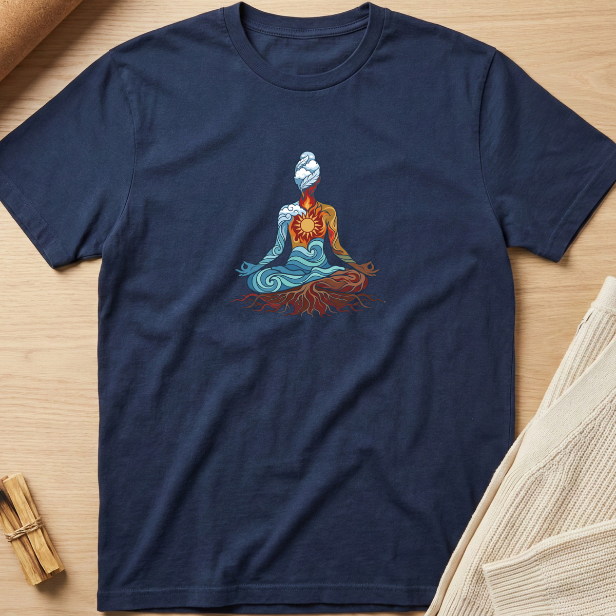A navy blue short-sleeve t-shirt laid flat on a light wood surface, featuring a colorful graphic of a meditating figure with blue, orange, and red details. The shirt is styled with a beige knit fabric and small wooden sticks in the corners. Minimalist yoga apparel by pemuyo.