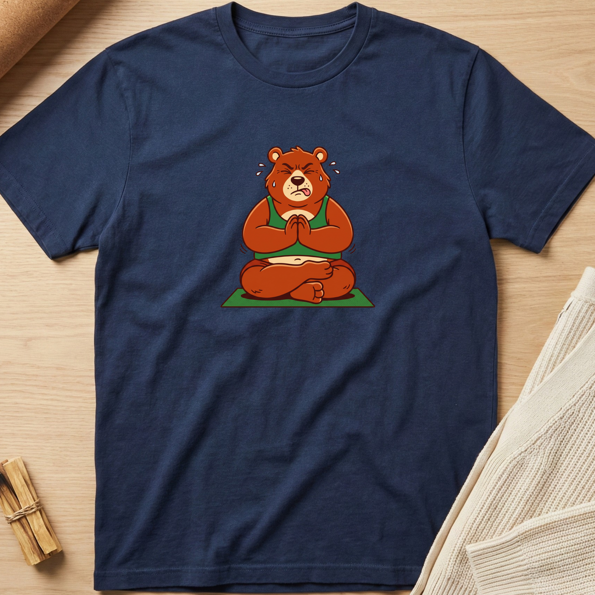 A navy blue crewneck t-shirt featuring a cartoon graphic of a brown bear meditating cross-legged on a green mat, laid flat on a light wood background with styled props. Minimalist yoga apparel by pemuyo.