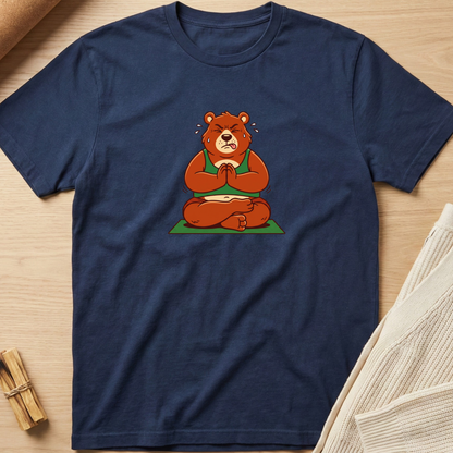 A navy blue crewneck t-shirt featuring a cartoon graphic of a brown bear meditating cross-legged on a green mat, laid flat on a light wood background with styled props. Minimalist yoga apparel by pemuyo.
