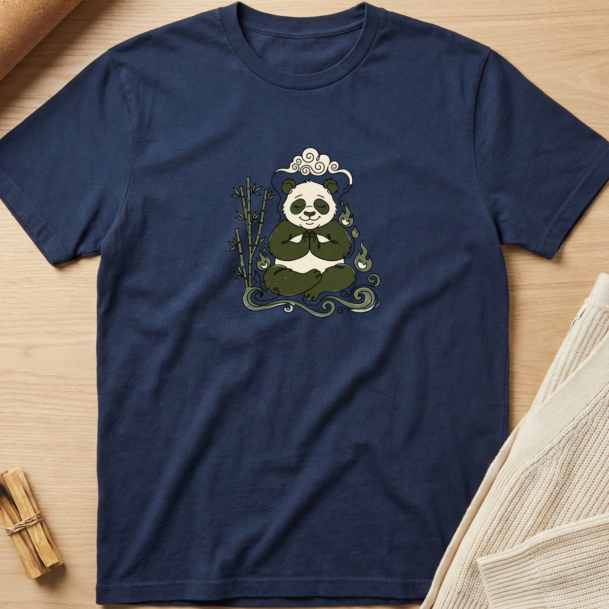 A navy blue short-sleeve t-shirt with a central graphic of a panda meditating on bamboo, styled flat on a light wood background with neutral props. Minimalist yoga apparel by pemuyo.