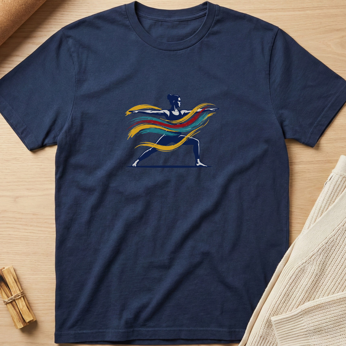 A navy blue short-sleeve t-shirt laid flat on a light wood surface, featuring a colorful graphic of a warrior silhouette with flowing yellow, orange, and red stripes. Styled flat-lay props include a rolled tan material in the top-left and a folded cream knit fabric in the bottom-right. Minimalist yoga apparel by pemuyo.