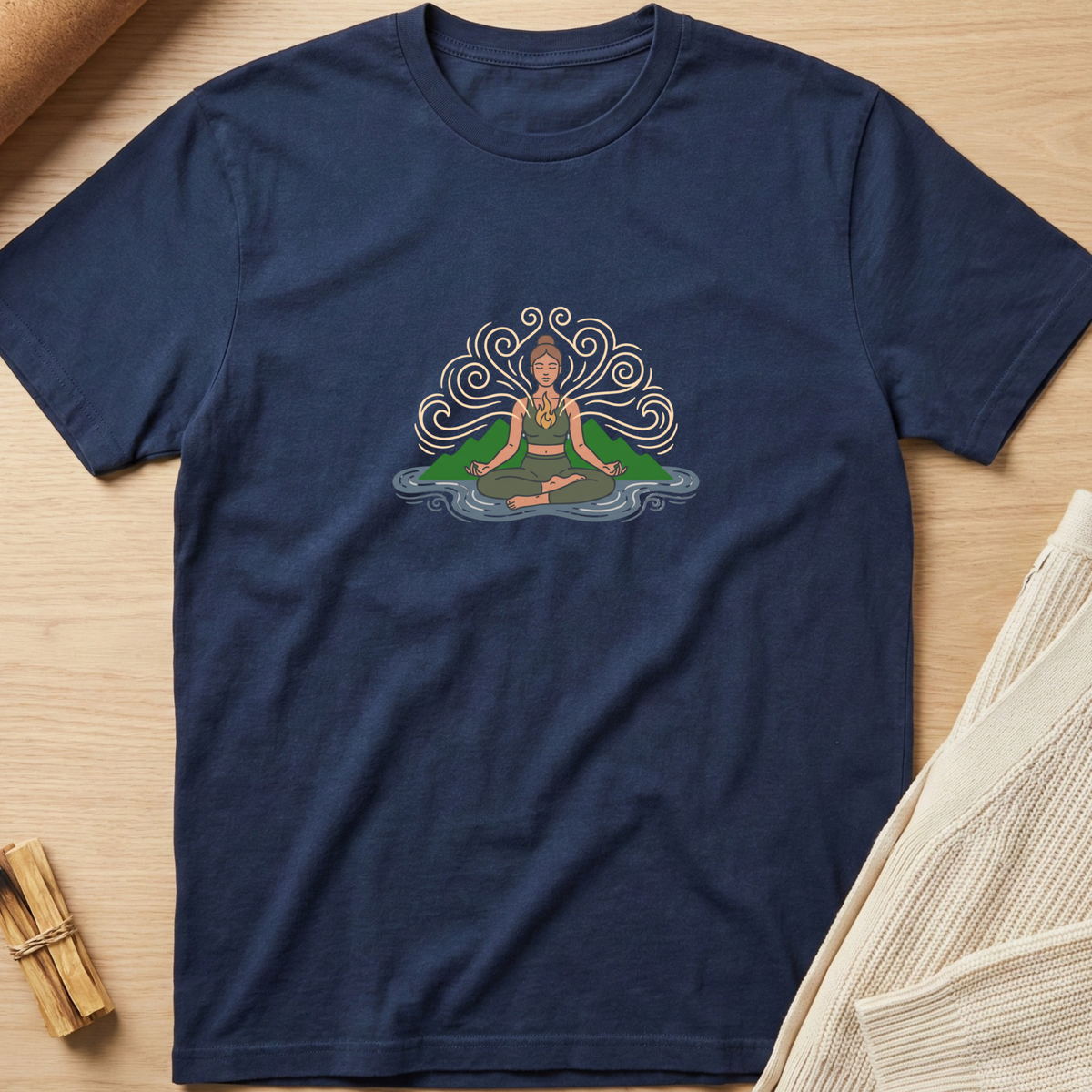 A navy blue short-sleeve t-shirt with a central graphic of a meditating figure surrounded by swirling patterns, laid flat on a light wood background with neutral props. Minimalist yoga apparel by pemuyo.