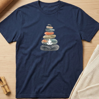 A navy blue short-sleeve t-shirt laid flat on a light wood surface, featuring a central graphic of stacked stones in muted earthy tones with a small white meditating figure at the base. A rolled tan fabric and a bundle of small wooden sticks are partially visible at the edges. Minimalist yoga apparel by pemuyo.