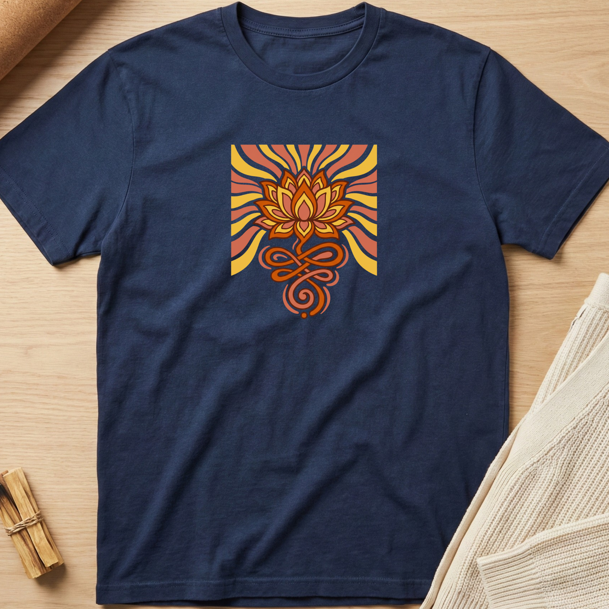 A navy blue short-sleeve t-shirt laid flat on a light wood surface, featuring a square graphic on the chest with a stylized orange and yellow lotus flower and swirling lines. Minimalist yoga apparel by pemuyo.