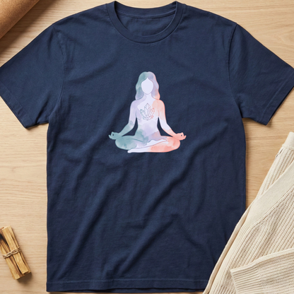 A navy blue short-sleeve t-shirt laid flat on a light wood surface, featuring a pastel watercolor-style graphic of a seated meditating figure on the chest. The shirt is styled with neutral props, including a beige knit fabric and a small bundle of wooden sticks. Minimalist yoga apparel by pemuyo.