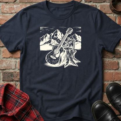Navy blue t-shirt with a white graphic of a guitar leaning against a tree stump with mountains in the background, displayed on a brick surface. Minimalist music apparel by pemuyo.