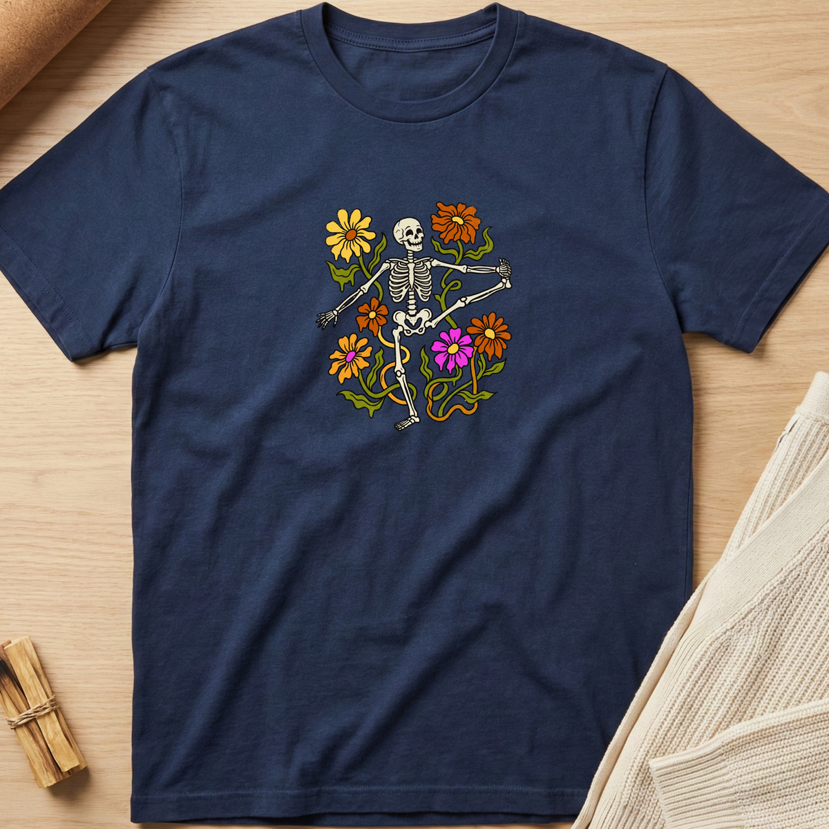 A navy blue short-sleeve t-shirt laid flat on a light wood surface, featuring a colorful graphic of a dancing skeleton surrounded by yellow, orange, and pink flowers. Styled flat-lay props include a beige knit fabric on the right and a small bundle of wooden sticks on the bottom left. Minimalist yoga apparel by pemuyo.