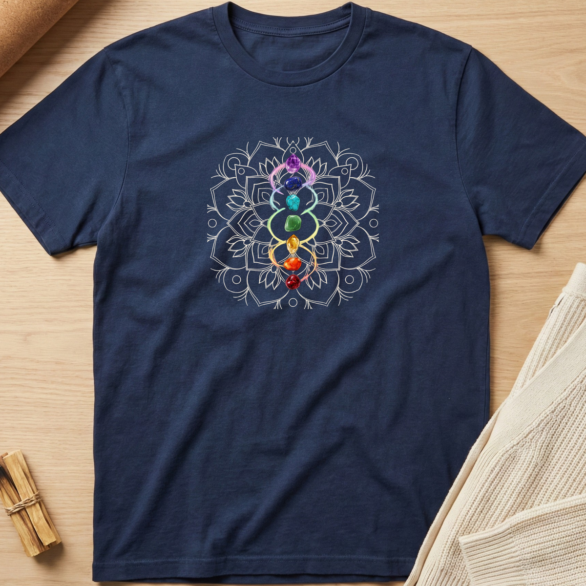 A navy blue short-sleeve t-shirt with a white mandala design and a vertical rainbow chakra symbol printed on the chest, displayed flat on a light wood background with neutral props. Minimalist yoga apparel by pemuyo.