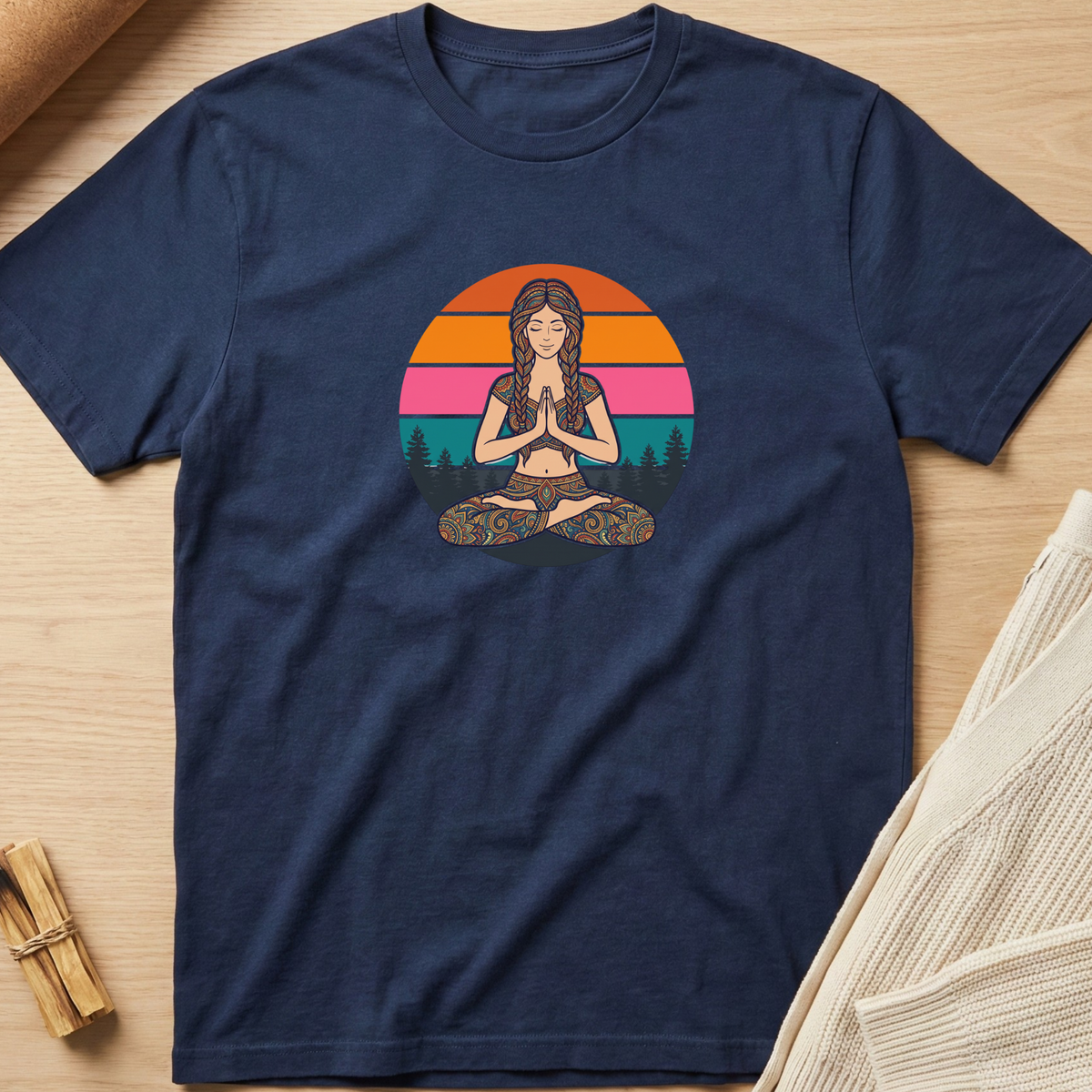 A navy blue short-sleeve t-shirt with a colorful retro-style graphic of a woman meditating in front of a sunset, laid flat on a light wood background with neutral props. Minimalist yoga apparel by pemuyo.