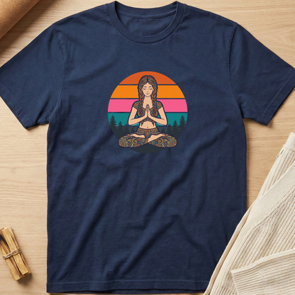 A navy blue short-sleeve t-shirt with a colorful retro-style graphic of a woman meditating in front of a sunset, laid flat on a light wood background with neutral props. Minimalist yoga apparel by pemuyo.