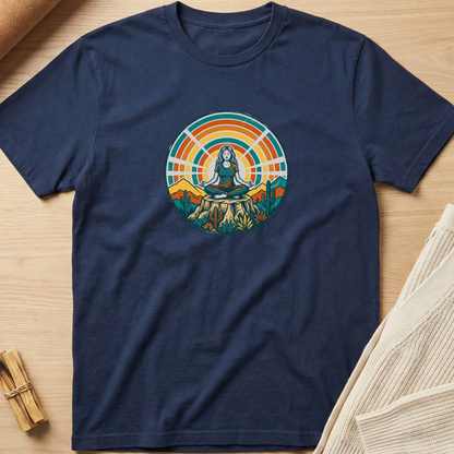 A navy blue short-sleeve t-shirt with a colorful retro circular graphic of a meditating figure on a mountain peak, laid flat on a light wood background with neutral props. Minimalist yoga apparel by pemuyo.