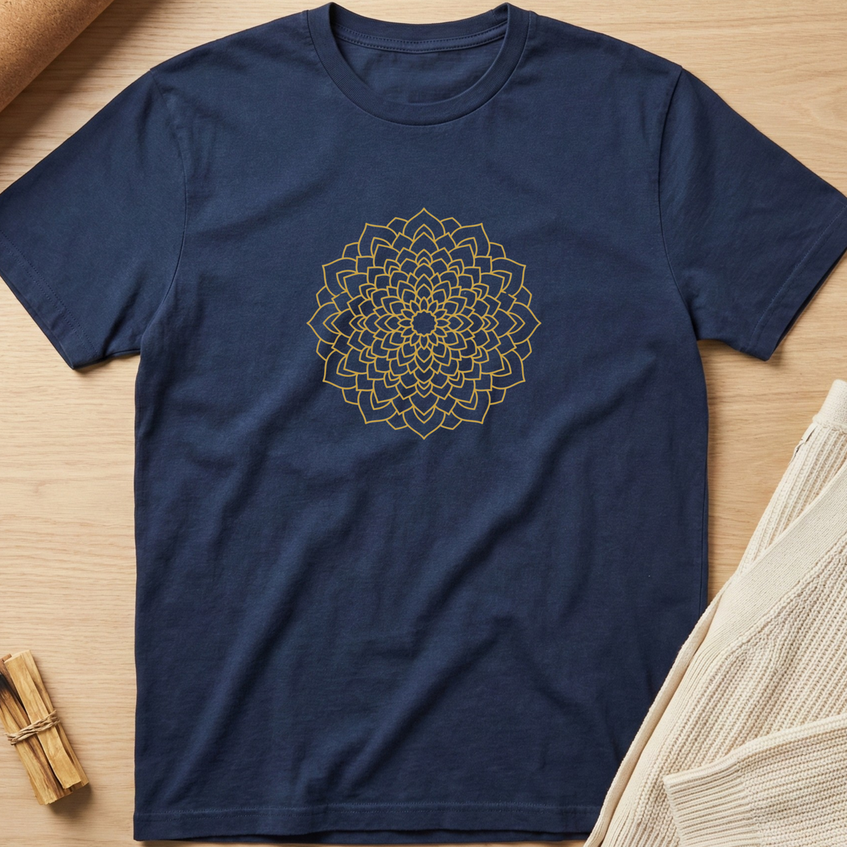 A navy blue short-sleeve crewneck t-shirt featuring a gold geometric mandala design printed on the chest, laid flat on a light wood background with neutral props. Minimalist yoga apparel by pemuyo.