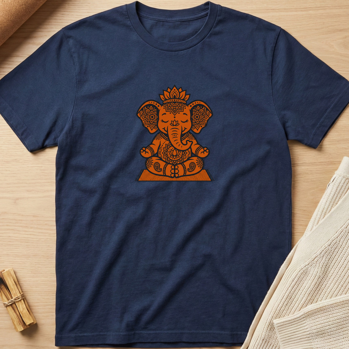 A navy blue short-sleeve t-shirt with a central orange Ganesha graphic, laid flat on a light wood background with neutral props. Minimalist yoga apparel by pemuyo.